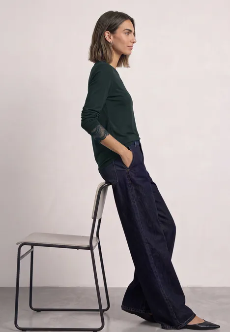 Langarm Shirt mit Rundhals und Spitze stormy green Eine Frau sitzt seitlich auf einem Hocker, trägt ein grünes Oberteil, weite blaue Hosen und flache Schuhe.