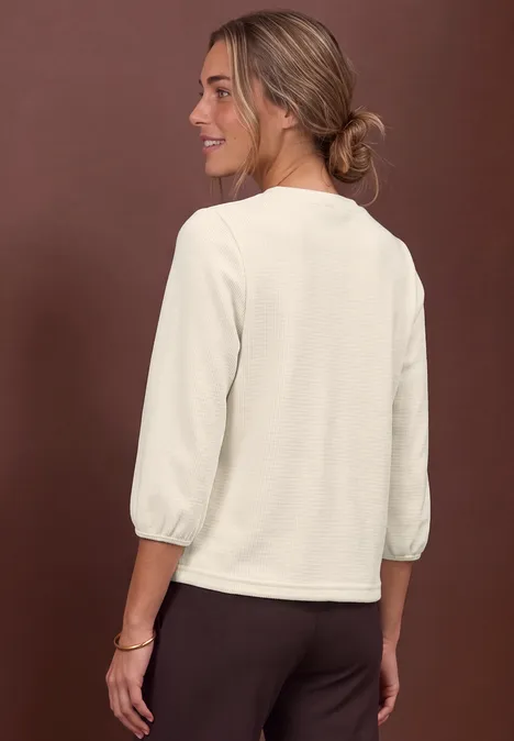 Frau von hinten seitlich in cremefarbener Bluse mit 3/4-Ärmeln und dunkler Hose vor braunem Hintergrund.