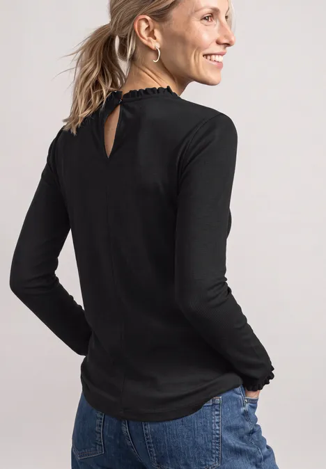 Frau in schwarzem Langarmshirt mit Rüschen am Kragen und Saum, blauen Jeans, im Profil lächelnd.