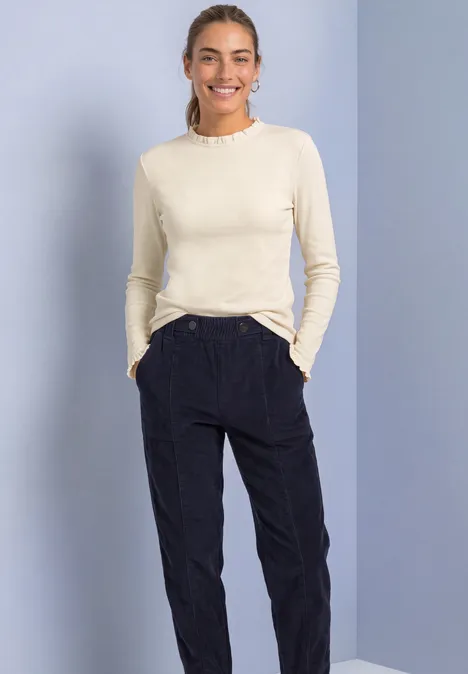 Vrouw lacht, draagt crèmekleurig lang shirt en zwarte broek, voor een lichtblauwe achtergrond.