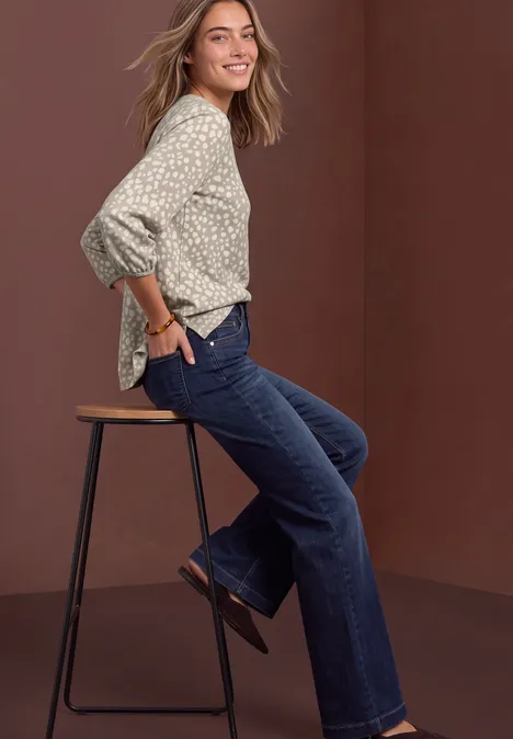Eine lächelnde Frau sitzt seitlich auf einem Barhocker, trägt geblümte Bluse, dunkle Jeans und Schuhe.