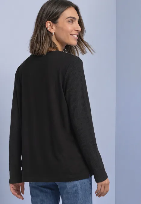T-shirt à manches longues texturé et boutonné Black Femme souriante regarde par-dessus son épaule, portant un pull noir et un jean devant un fond clair.