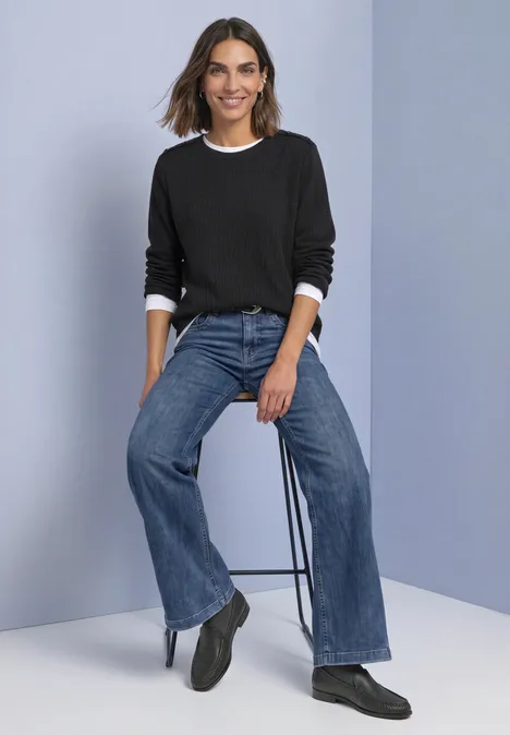 T-shirt à manches longues texturé et boutonné Black Une femme assise sur un tabouret porte un pull noir à col blanc, un jean large bleu et des bottines noires.
