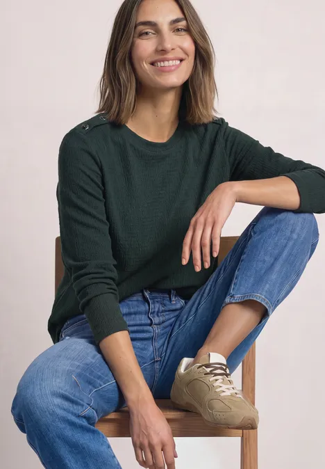 Een glimlachende vrouw in een groene trui en spijkerbroek zit ontspannen op een houten stoel.