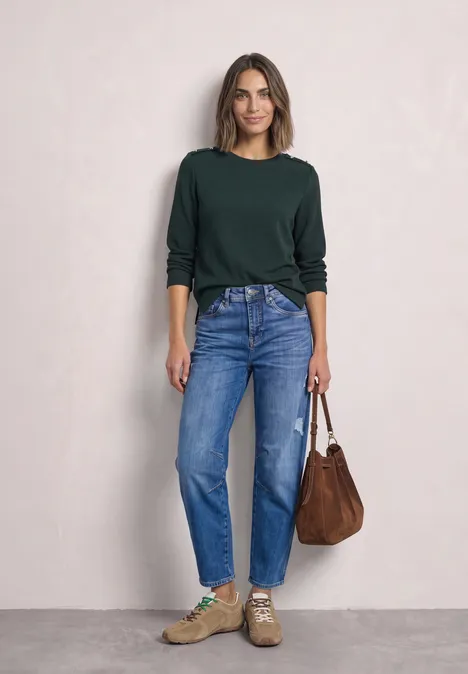 Lachende vrouw voor lichte muur in groene trui, blauwe jeans, sneakers en een bruine schoudertas.