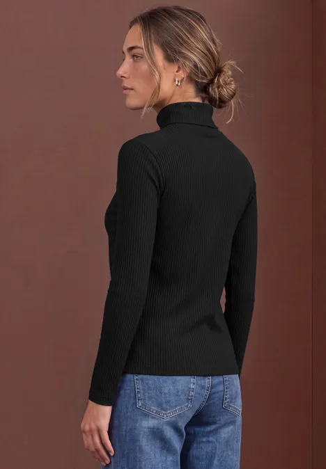 Eine Frau in schwarzem Rollkragenpullover und Jeans steht seitlich vor braunem Hintergrund.