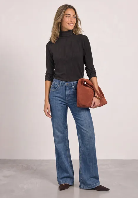 Vrouw in zwarte coltrui, wijde jeans en zwarte schoenen draagt een bruine tas voor een lichte achtergrond.