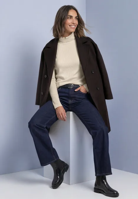 T-shirt basique uni à col roulé pure beige Femme en manteau marron, pull col roulé crème, jean large et bottines noires, assise souriante sur un bloc blanc