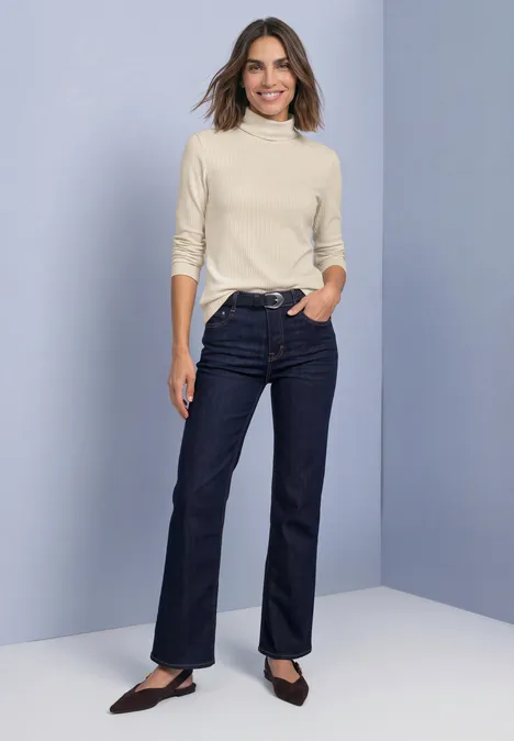 T-shirt basique uni à col roulé pure beige Femme portant un pull à col roulé clair, un jean foncé et des chaussures plates noires devant un fond uni