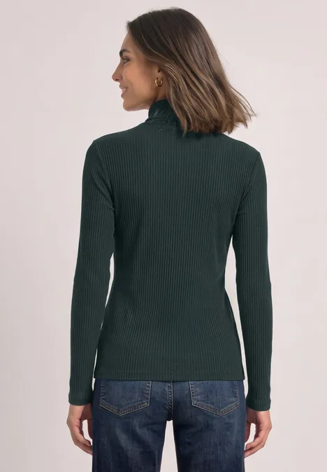 Rückansicht einer Frau in dunkelgrünem Rollkragenpullover und blauen Jeans vor hellem Hintergrund.