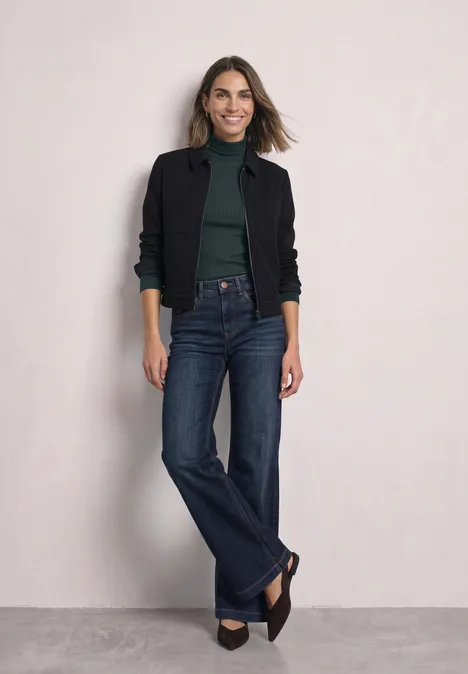 Eine Frau in schwarzer Jacke, grünem Oberteil und weiten Jeans steht vor hellem Hintergrund und lächelt.