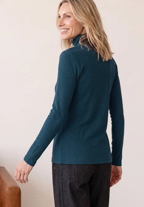 Vrouw in donkergroene coltrui en grijze broek, kijkt over haar schouder, glimlachend.