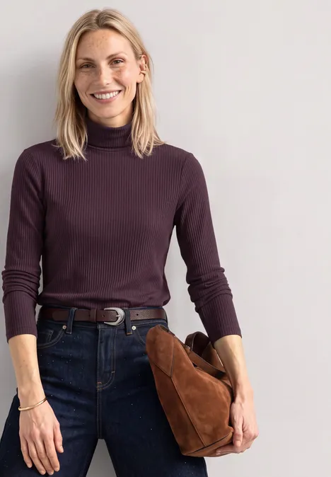 Frau mit blondem Haar, dunkelviolettem Rollkragenpullover und Jeans, hält eine braune Tasche und lächelt.