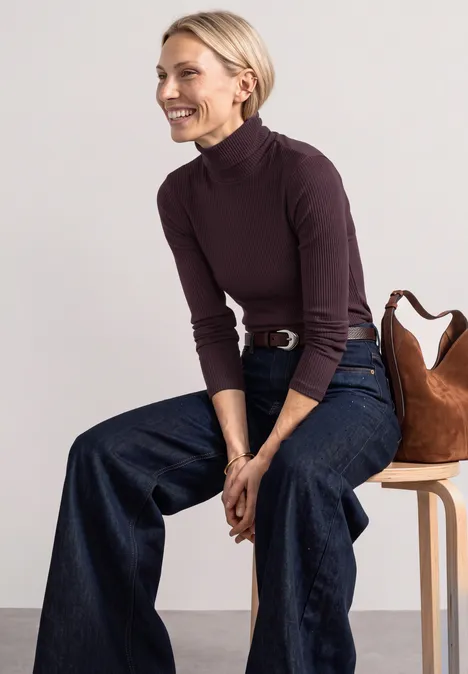 Frau in lila Rollkragenpullover und weiten Jeans sitzt lächelnd auf einem Hocker mit Tasche.
