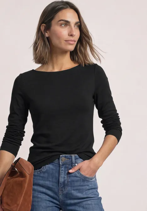 T-shirt basique à col bateau Black Frau in schwarzem Langarmshirt, blauen Jeans und brauner Wildledertasche vor hellem Hintergrund.