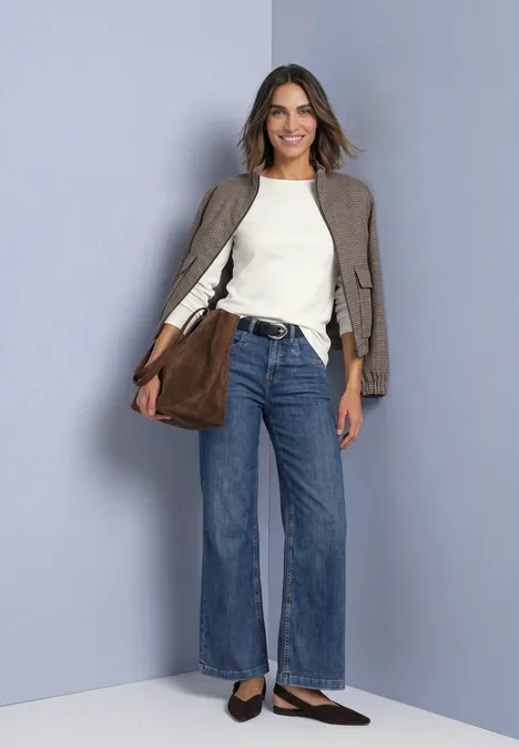 Femme souriante en jean évasé, top blanc, veste taupe et sac marron devant un fond gris.
