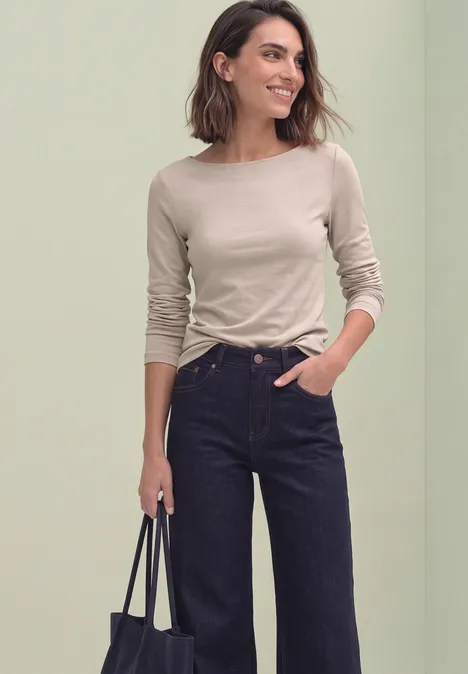 T-shirt basique à col bateau clay sand Eine lächelnde Frau in beigefarbenem Langarmshirt, dunklen Jeans mit weitem Bein und schwarzer Tasche