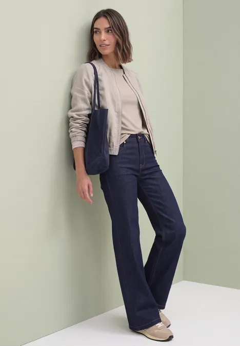 T-shirt basique à col bateau clay sand Frau in beigem Pullover, Blazer, weiter Jeans und Tasche lehnt an hellgrüner Wand.