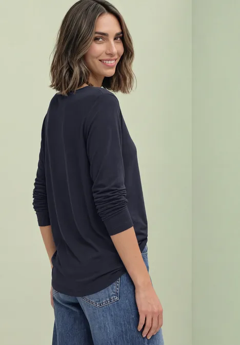 Basic Shirt mit U-Boot-Ausschnitt indigo night Eine lächelnde Frau blickt über die Schulter, trägt ein dunkelblaues Shirt und steht vor hellem Hintergrund.