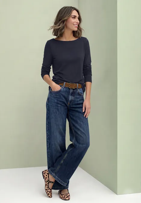 Basic Shirt mit U-Boot-Ausschnitt indigo night Eine Frau lehnt an einer Wand, trägt schwarzen Pullover und weite Jeans mit Leoparden-Gürtel.