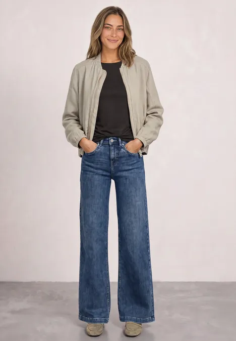 Basic Shirt mit U-Boot-Ausschnitt black coffee Frau mit beige Bomberjacke, schwarzem Shirt und weiten Blue-Jeans vor schlichter Wand.