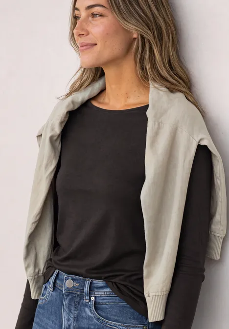 Basic Shirt mit U-Boot-Ausschnitt black coffee Frau trägt schwarzen Langarmshirt und leichte Jacke über den Schultern, steht lächelnd vor hellem Hintergrund.