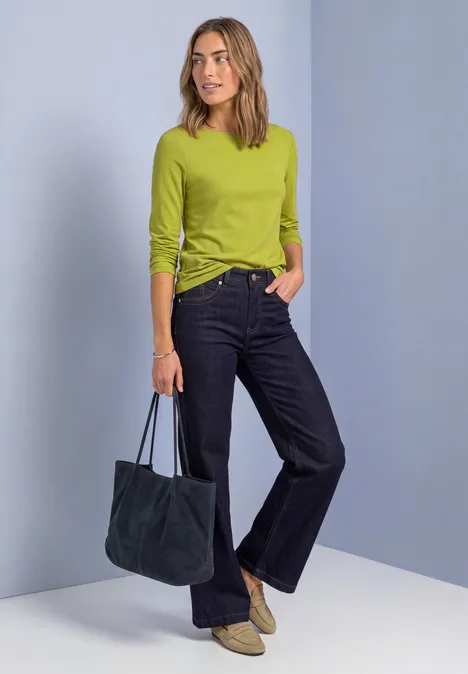 T-shirt basique à col bateau et manches longues apple lime Frau in limonengrünem Langarmshirt, dunklen Jeans und beigen Boots, mit dunkelblauer Tasche, vor hellblauem Hintergrund.