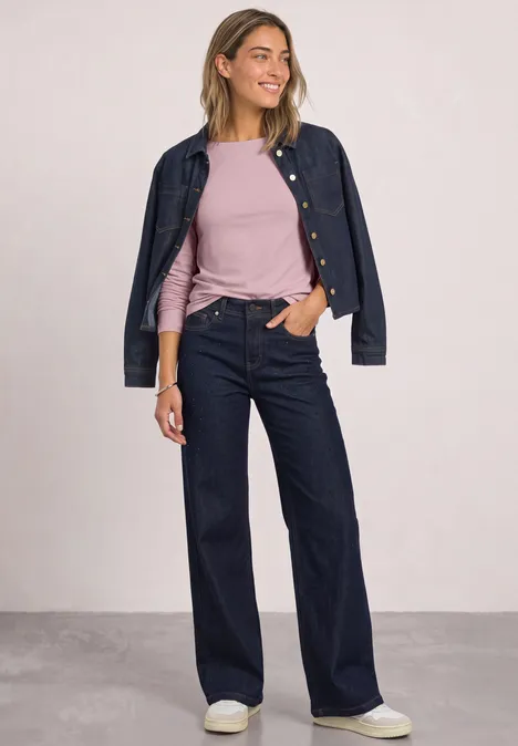 T-shirt basique à col bateau et manches longues quiet rose Frau in dunkler Jeansjacke und -hose, rosa Shirt und weißen Sneakers, steht entspannt mit einer Hand in der Tasche.