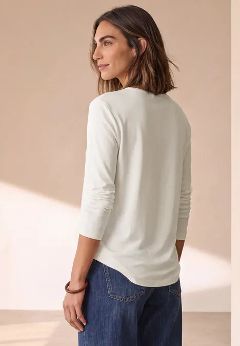 Vrouw in wit shirt met lange mouwen en spijkerbroek, kijkt ontspannen opzij.