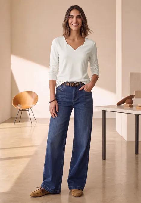 Vrouw in lichte kamer met witte V-hals top, brede blauwe jeans en bruine loafers, casual en stijlvol.
