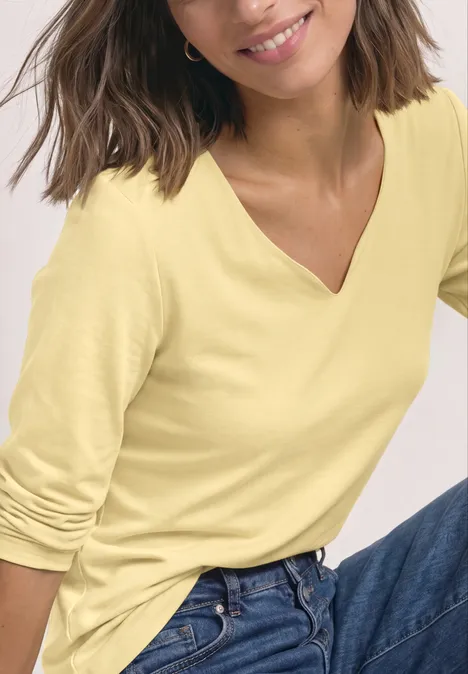 Langarm Shirt mit Herzausschnitt sunbeam yellow Frau mit gelbem V-Ausschnitt-Shirt und blauer Jeans, sitzt lächelnd.