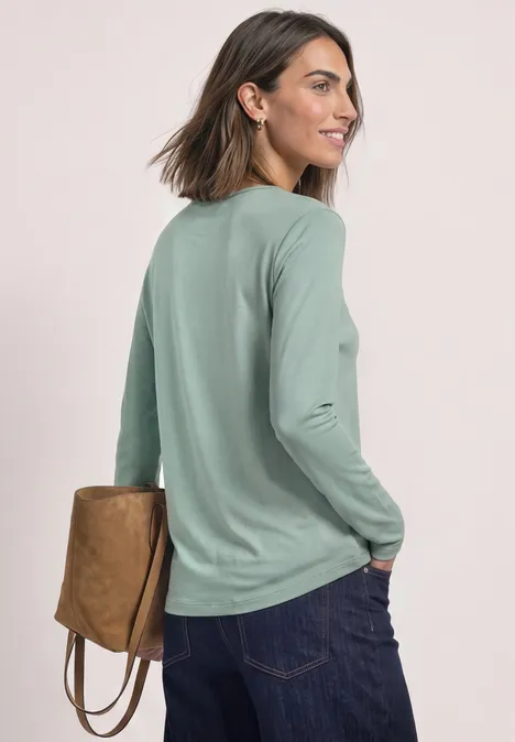 Vrouw met kort bruin haar, gezien van achter, draagt een groen langarmshirt, donkere spijkerbroek en een bruine tas.