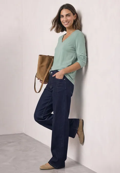 Een vrouw leunt tegen een muur, draagt een lichtgroene trui, brede jeans en bruine enkellaarsjes.