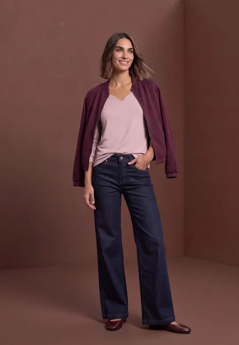 Eine lächelnde Frau in violetter Bomberjacke, rosa Top und weiten, hochgeschnittenen Jeans vor braunem Hintergrund.