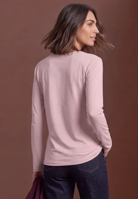 Frau in rosa Langarmshirt und Jeans, leicht von hinten, Blick über die Schulter vor neutralem Hintergrund.