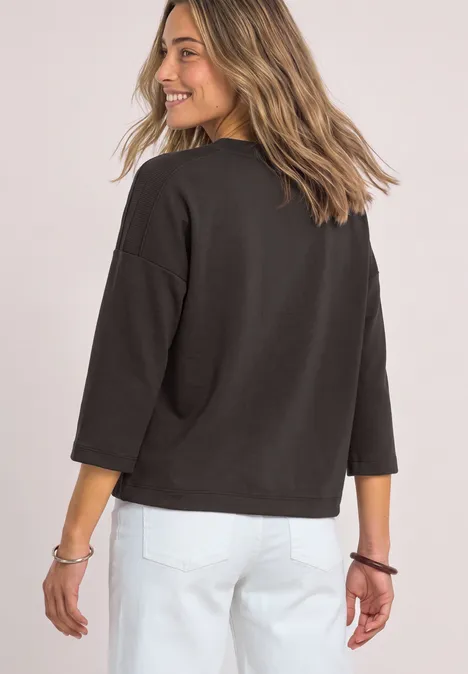 3/4-Arm Shirt mit Boat Neck und Tunnelzug black coffee Frau in schwarzem Pullover und weißer Hose blickt lächelnd über die Schulter.
