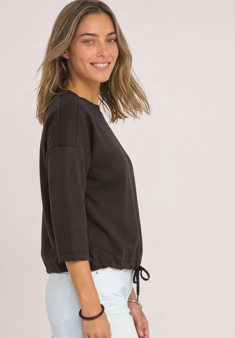 3/4-Arm Shirt mit Boat Neck und Tunnelzug black coffee Frau im Profil lacht, trägt schwarzes 3/4-Arm-Shirt mit Kordelzug und weiße Hose vor hellem Hintergrund.