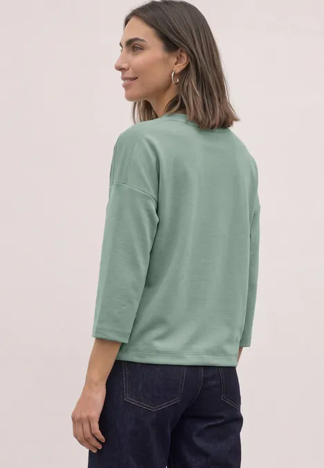 3/4-Arm Shirt mit Boat Neck und Tunnelzug eucalyptus green Frau im Profil mit lockerem grünen 3/4-Arm-Shirt und dunkler Jeans vor neutralem Hintergrund.