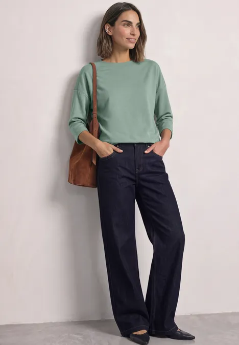 3/4-Arm Shirt mit Boat Neck und Tunnelzug eucalyptus green Frau vor hellem Hintergrund in grünem Pullover, weiten Jeans und brauner Schultertasche.