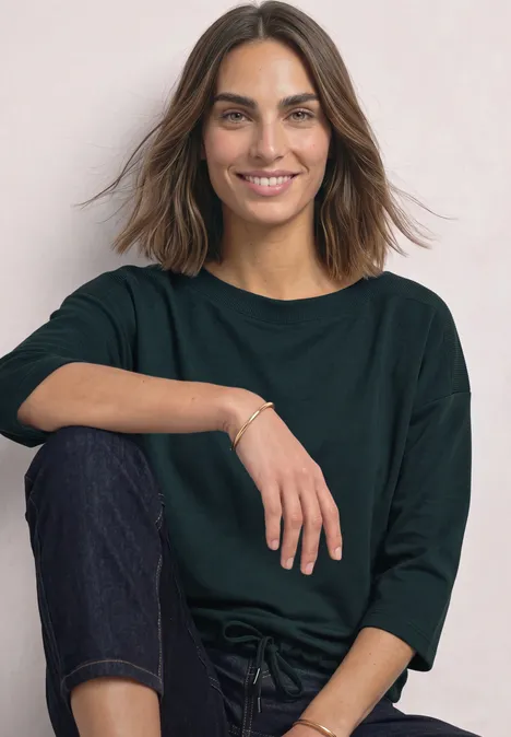 3/4-Arm Shirt mit Boat Neck und Tunnelzug stormy green Eine lächelnde Frau in grünem Oberteil und Jeans sitzt entspannt vor hellem Hintergrund.