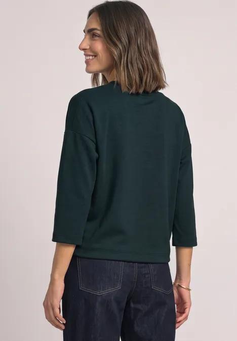 3/4-Arm Shirt mit Boat Neck und Tunnelzug stormy green Frau von hinten in dunkelgrünem Pullover mit 3/4-Ärmeln und Jeans vor neutralem hellen Hintergrund.