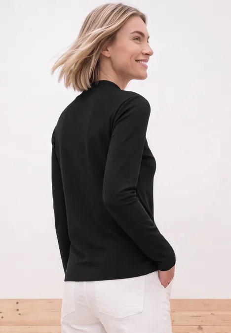 Een vrouw staat zijwaarts en lacht, gekleed in een zwarte lange mouw top en witte jeans.