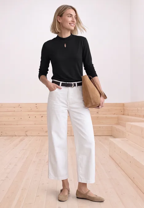 Een lachende vrouw draagt een zwarte trui, witte wijde broek, bruine loafers en draagt een leren tas.