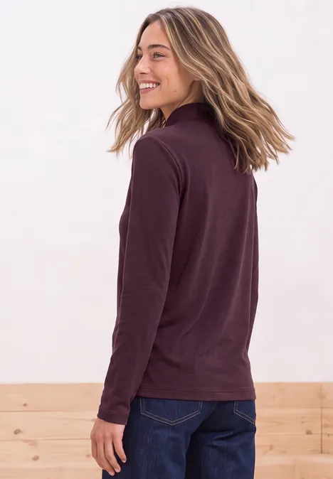 Een glimlachende vrouw in profiel met bordeauxrode trui en spijkerbroek, met los blond haar.