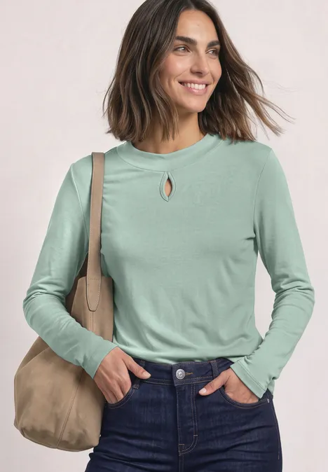 Lächelnde Frau in hellgrünem Langarmshirt mit Schlitzdetail, Jeans und beiger Umhängetasche vor hellem Hintergrund.