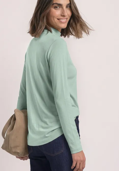 Frau im Profil trägt hellgrünes Langarmshirt, dunkle Jeans, hält eine beige Tasche und lächelt.
