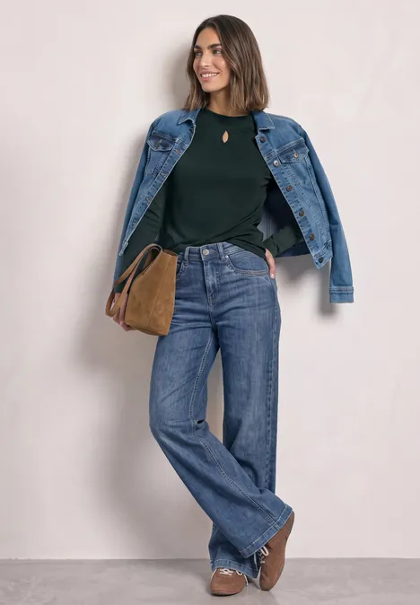 Frau mit Jeansjacke über den Schultern, grünem Pullover, weiten Jeans, braunen Stiefeln und Umhängetasche.