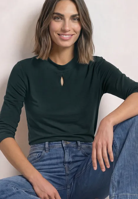 Eine lächelnde Frau in dunkelgrünem Langarmshirt mit kleinem Ausschnitt und blauen Jeans sitzt locker.