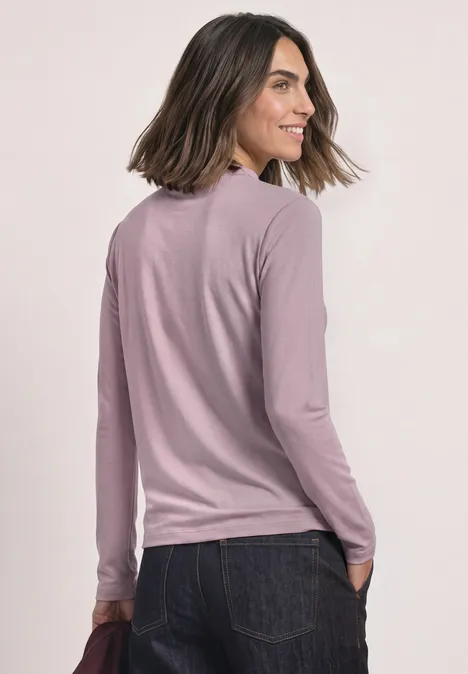 Vrouw in een roze langärmelig shirt en spijkerbroek, kijkt over haar schouder en glimlacht.