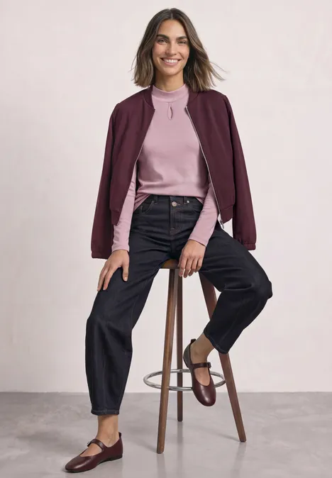Vrouw zit op kruk, draagt roze trui, bordeaux jasje, zwarte jeans en bruine loafers.